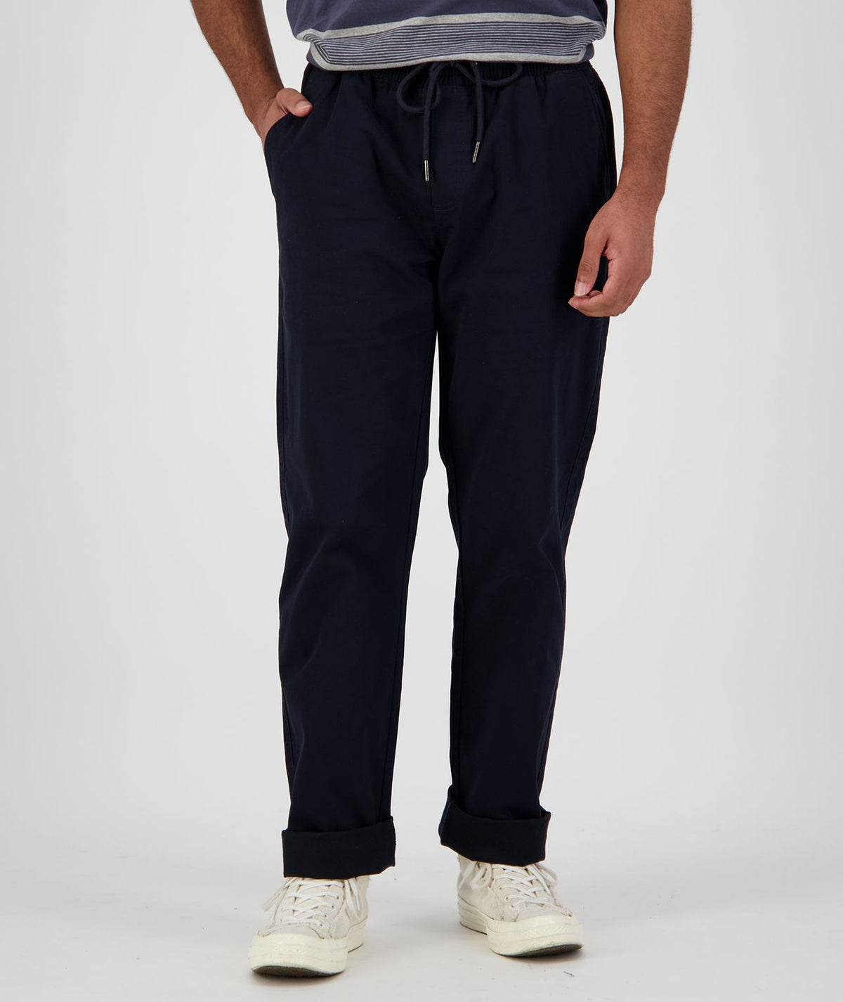 A person stands wearing the Swanndri Karaka Pant in dark blue and white sneakers. The individual has their hands in the pants' pockets and is seen from the shoulders down against a plain, light-colored background.