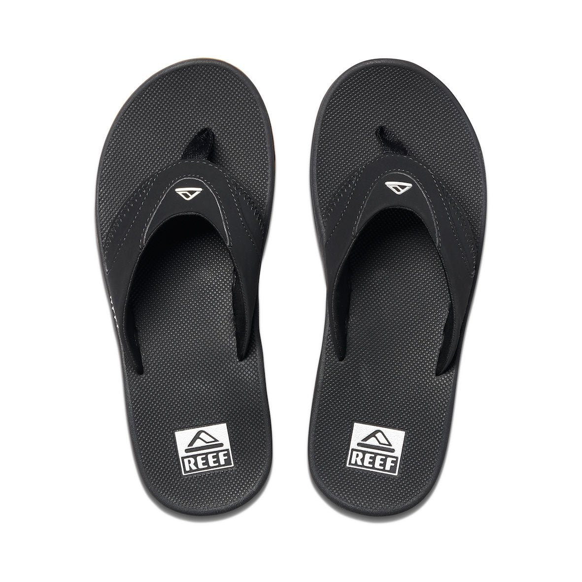 The REEF Fanning flip-flops are black with textured soles and feature a stylized wave logo on the synthetic strap. The brand name "REEF" is prominently displayed on the heel, and they include an EVA footbed for added comfort.