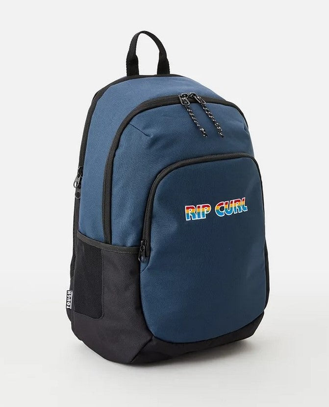 A Rip Curl Ozone 30L Icons Eco Backpack in blue and black, featuring a colorful "RIP CURL" logo on the front pocket. Made from recycled plastic, it includes multiple zippered compartments and a small black handle on top.
