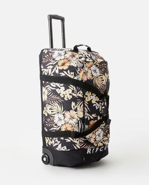 The Rip Curl Jupiter 80L Paradise Travel Bag features a floral-patterned design with yellow, white, and beige flowers accompanied by green leaves on a black background. It includes an extended handle for ease of transport and has a small tag at the bottom displaying the brand name "RIP CURL.