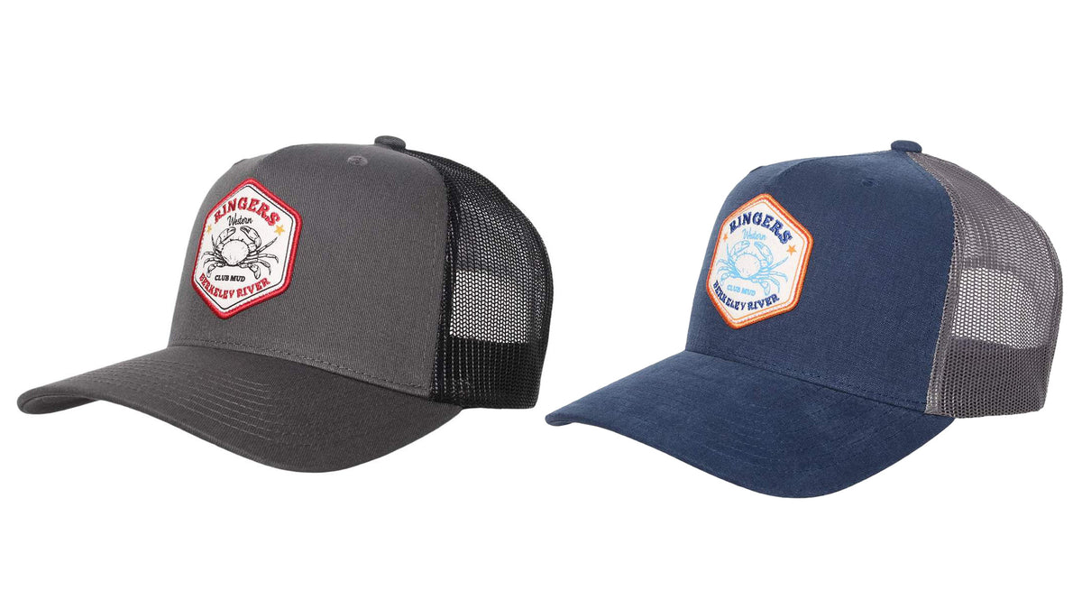 Two baseball caps are displayed side by side. The left cap is gray with a black mesh back, featuring a Ringers Western patch with a red border and logo. The right cap, known as the Ringers Western Club Mud Trucker Cap, is navy blue with a gray mesh back and features a patch with a blue border and logo.