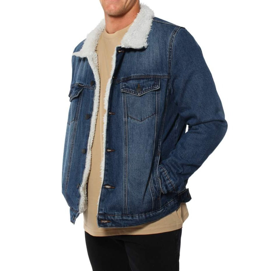A person wearing a Ringers Western Duncan Denim Jacket with a cozy sherpa lining. The jacket is open to show a beige shirt underneath. The person stands with one hand in their pocket and the other at their side, against a plain white background.