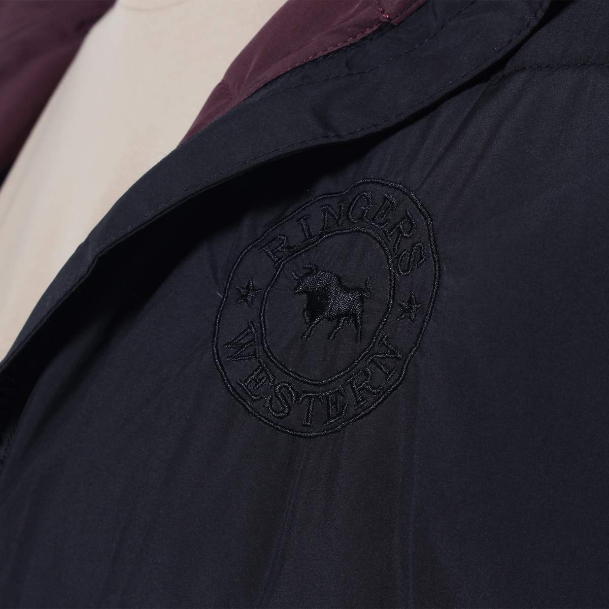 Close-up image of a dark-colored Ringers Western Bairnsdale Puffer Jacket displaying an embroidered circular logo that reads "RINGERS WESTERN" with a cow silhouette in the center and stars on either side. The jacket features a detachable hood and appears to be worn over a light-colored inner layer.
