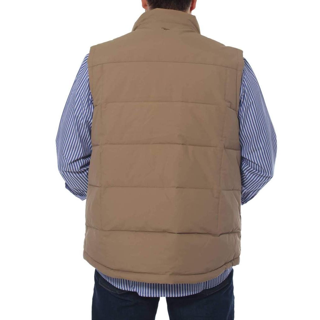 A person is seen from the back wearing a tan-colored Ringers Western Horsham Puffer Vest with a soft fleece lining over a blue and white striped shirt. The person has their hands in the pockets of the comfortable vest, and the bottom portion of dark blue jeans is visible. The background is plain and white.