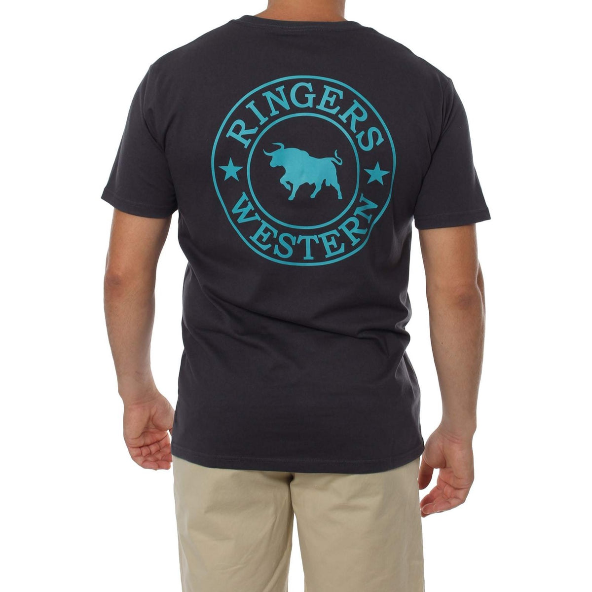 A person stands facing away, wearing the comfortable Ringers Western Signature Bull Classic T-Shirt, a dark crew neck with a turquoise circle logo on the back that features a bull silhouette in the center and "RINGERS WESTERN" along the top and bottom. They are also dressed in light beige shorts.