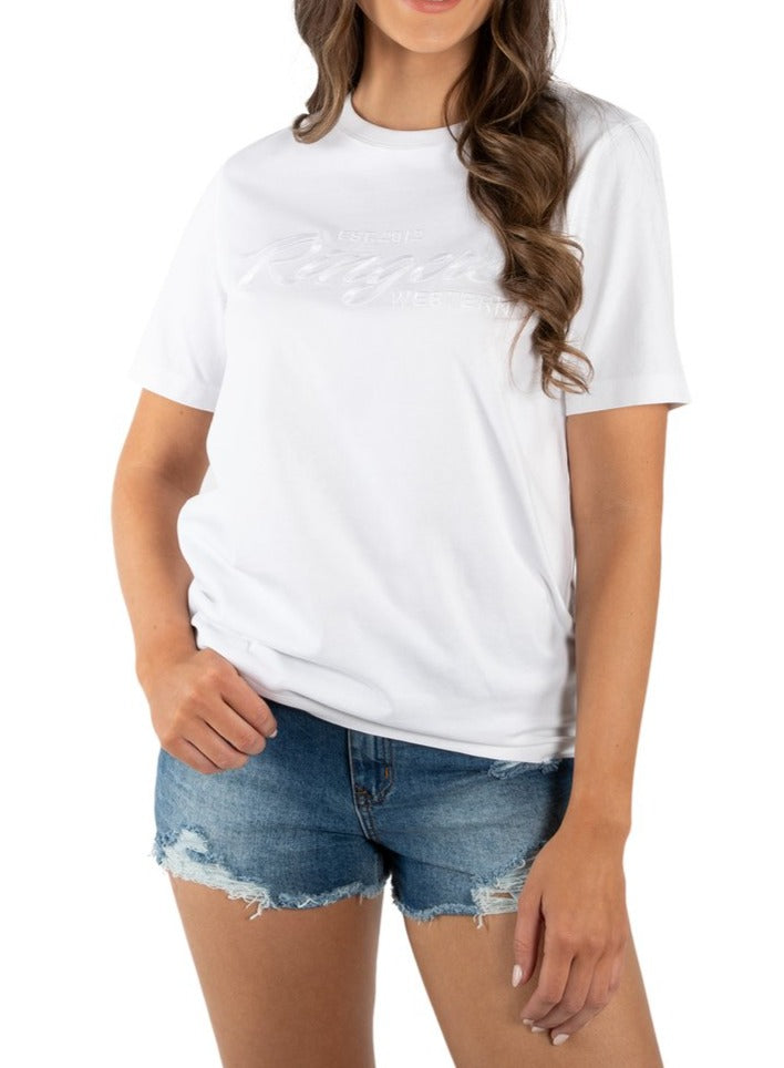 A person in a Ringers Western Women's Columbia Emboss Logo T-Shirt with denim shorts stands against a plain background. Their arms rest at their sides, and subtle writing on the 100% cotton shirt adds detail. Their long, wavy hair cascades down.