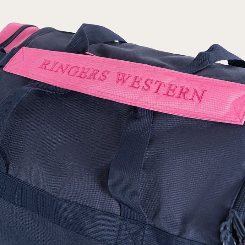 Close-up of a dark blue Ringers Western Rider Sports Bag featuring a pink handle with "RINGERS WESTERN" embroidered in capital letters. Several straps are visible around the bag, highlighting its design and durability, making it an ideal sports bag. The background is plain and neutral.