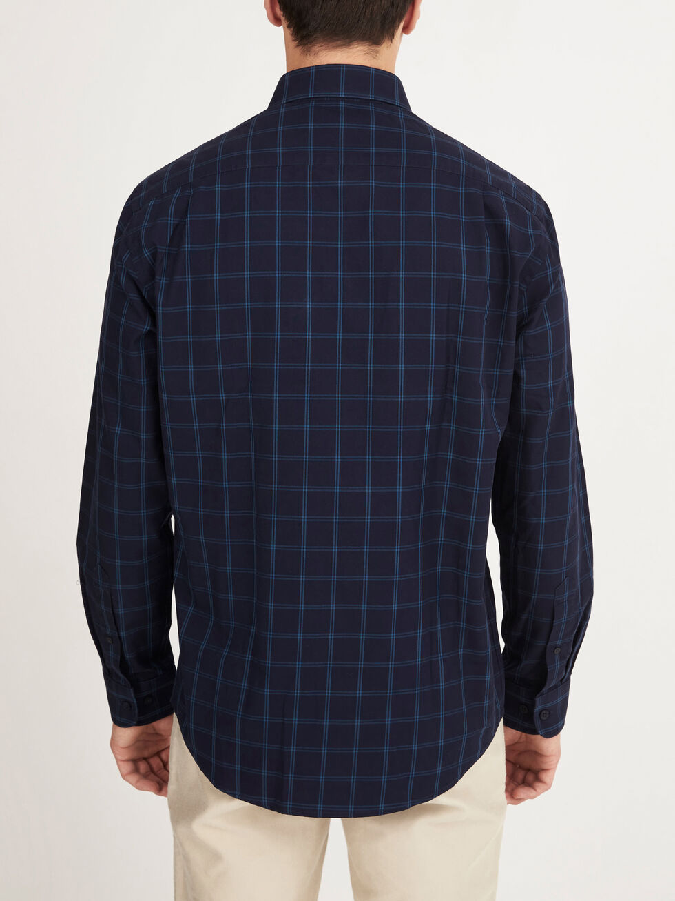 A man is shown from the back wearing the RM Williams Collins Shirt, a navy blue, long-sleeved, 100% cotton button-up with a light blue checkered pattern, paired with light-colored pants.
