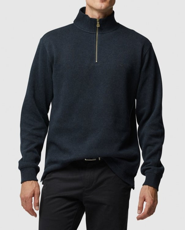 The man is dressed in a Rodd & Gunn Alton Ave Sweat, a dark half-zip design with a high collar. This stylish piece is elegantly matched with dark pants against a plain gray background.