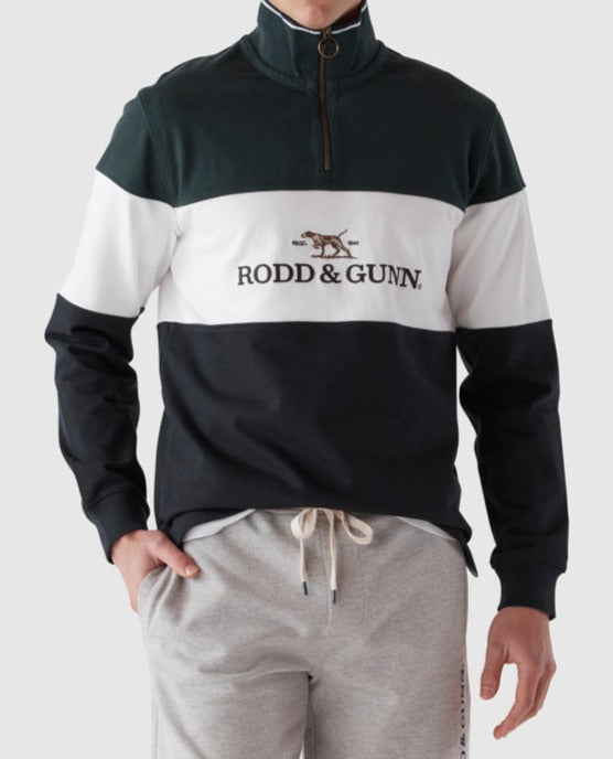 A person wearing the Rodd & Gunn Foresters Peak Sweat, a quarter-zip pullover in black, white, and dark green with "RODD & GUNN" and a dog logo embroidered on the chest. The 100% cotton top is perfect for casual wear. The person also sports light gray sweatpants with a drawstring waistband. The background is plain and neutral.