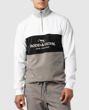 A person is wearing the stylish Rodd & Gunn Hillop Sweat, a 100% cotton jumper in white, grey, and black with a quarter-zip neckline and "Rodd & Gunn, New Zealand" printed in the center. They are also sporting matching grey pants with a drawstring waist. The background is plain grey.