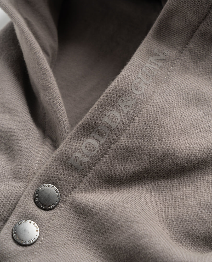 Close-up of the Rodd & Gunn Worser Bay Sweat, a light brown, comfortable hoodie with silver buttons and "FOOD & GUN" embroidered along the seam in a matching color. The high-quality soft fleece lining fabric ensures comfort. The buttons, engraved with "FOOD & GUN" around their edges, add a touch of subtle elegance to this piece.