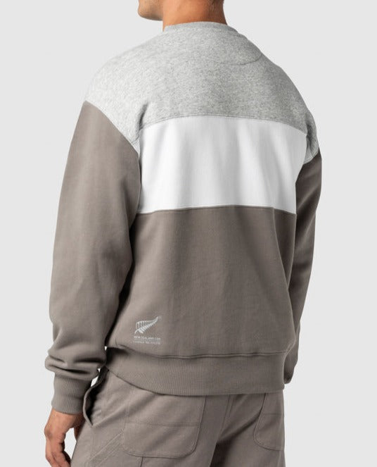 A person is wearing the Rodd & Gunn Mount Wesley Sweat, a casual color-blocked sweatshirt in gray, white, and light brown. Viewed from the back, it’s paired with light brown pants. The New Zealand national rugby team logo is on the lower left, and it features a cozy fleece lining for comfort.