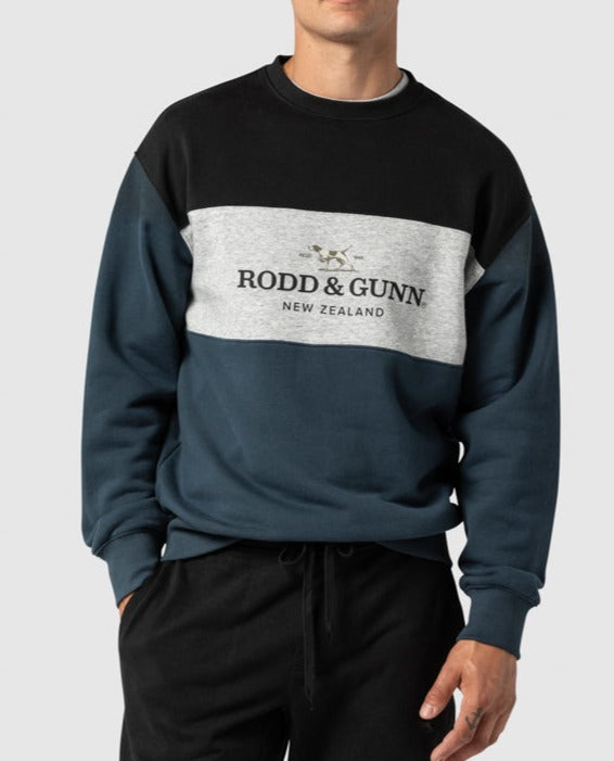 A man wearing the Rodd & Gunn Mount Wesley Sweat, a color-blocked crewneck sweatshirt with "RODD & GUNN NEW ZEALAND" text and logo on the chest. The comfortable jumper features black, grey, and dark teal sections, along with a soft fleece lining. The man stands with his hand in one pocket, against a plain background.