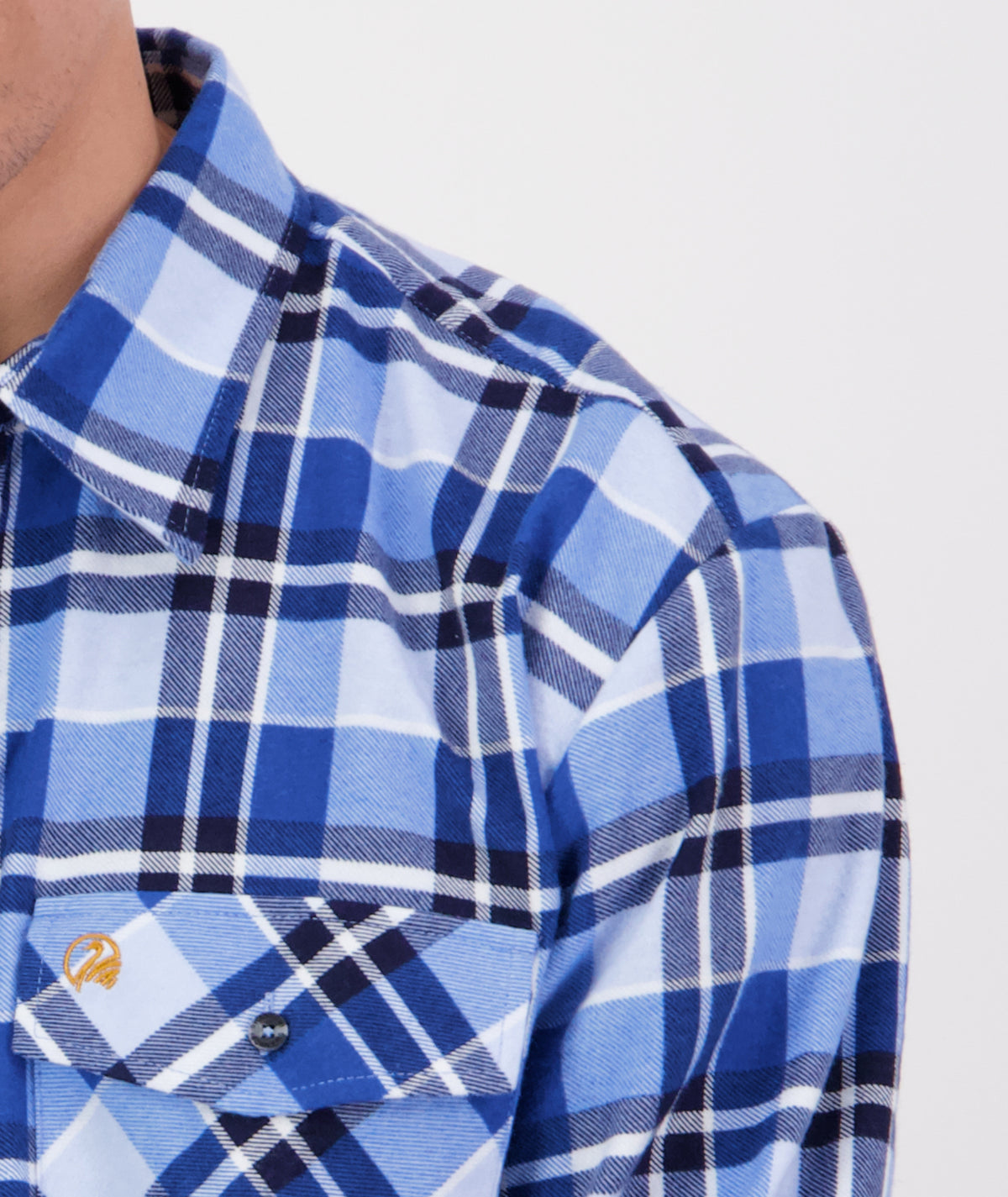 Close-up of a person wearing a Swanndri Egmont Flannelette Twin Pack blue and white plaid shirt made from brushed cotton, showcasing the collar, chest pocket, and button details. The focus is on the upper portion of the long sleeve shirt, with a small portion of the person's face partially visible on the left edge of the image.