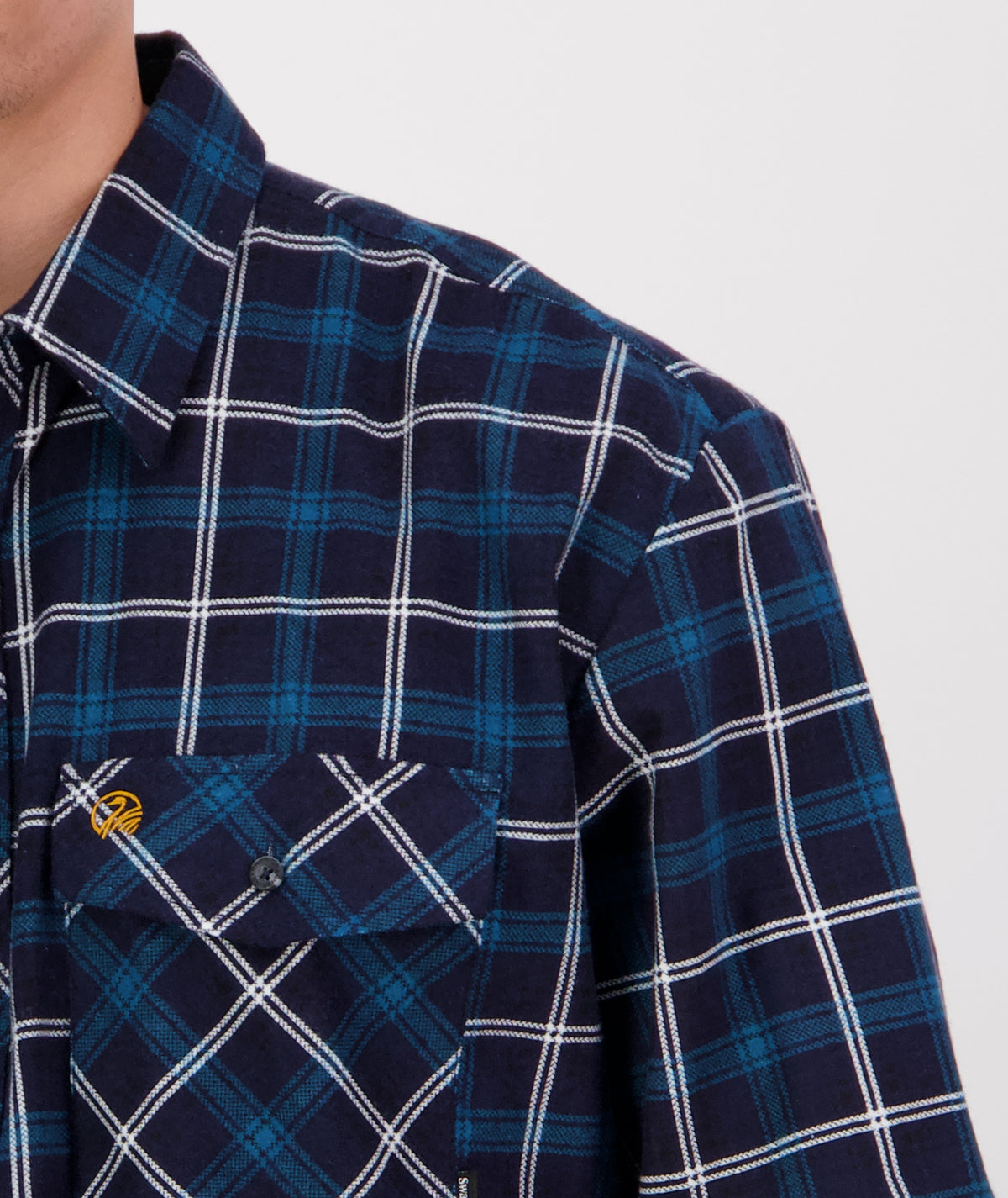 A close-up view of a person wearing a blue and white plaid long sleeve Swanndri Egmont Flannelette shirt made of brushed cotton. The shirt features a collar and a pocket with a subtle logo over the chest. Only the upper half body and part of the chest area are visible against a plain background.