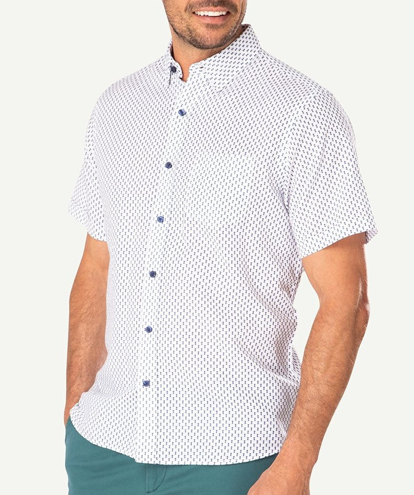 A person is wearing a Gazman Linen Blend Pineapple Print Shirt, featuring short sleeves, a white base with a small blue pattern, and blue buttons. The comfortable shirt, made of breathable fabric, pairs well with green trousers. With their right hand in their pocket, they stand against a plain light background.
