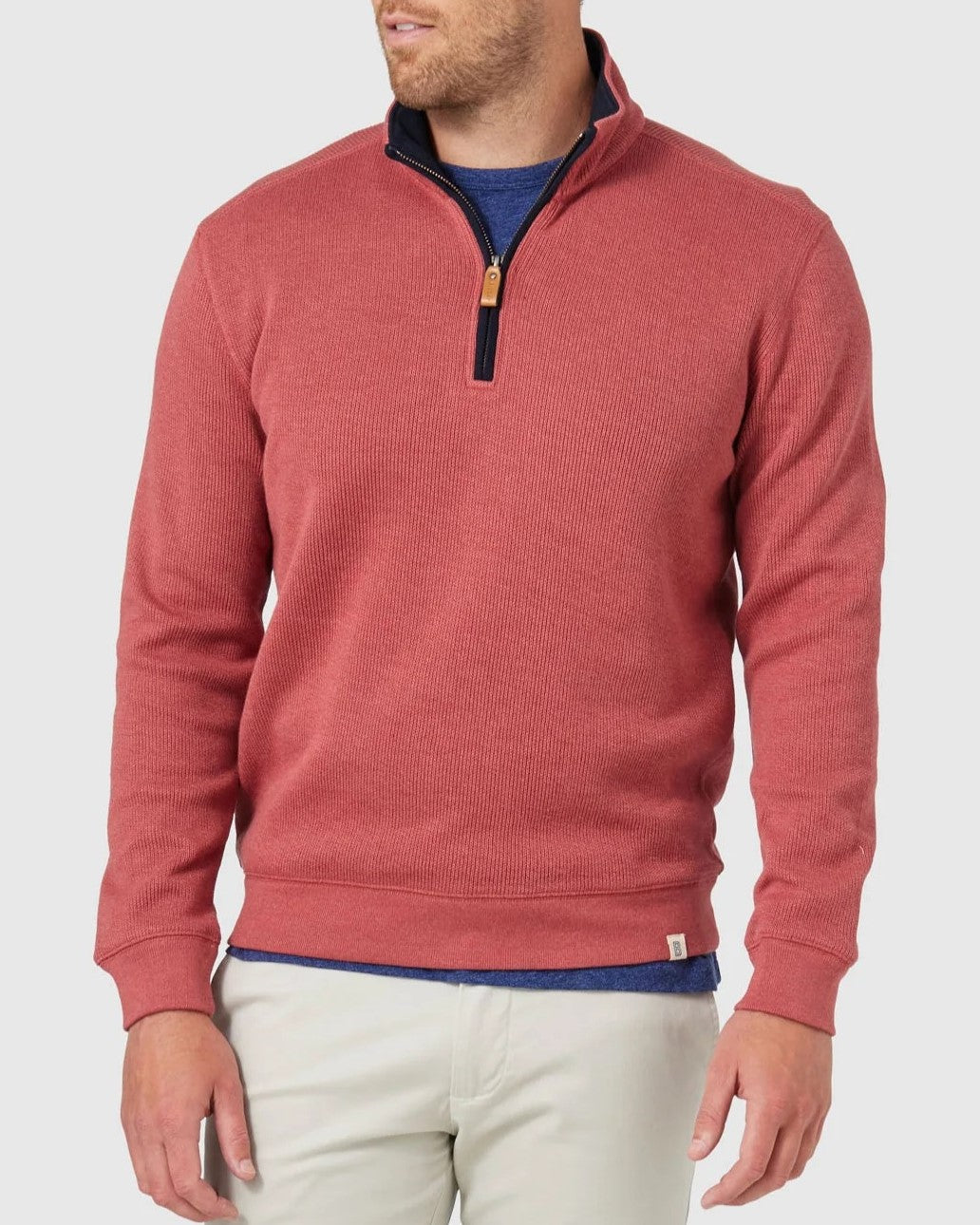 A man stands against a light gray background wearing the Blazer Heather Half Zip Sweat in red over a blue shirt and beige pants, his head slightly turned and hands relaxed at his sides.