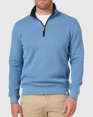 Wearing a light blue Blazer Heather Half Zip Sweat with a black collar and tan zipper pull, paired with beige pants creates casual sophistication. The plain, light-colored background enhances the ensemble's relaxed vibe effortlessly.