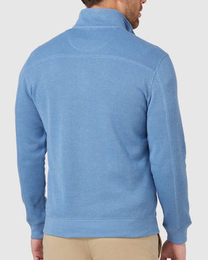 A person wearing the Blazer Heather Half Zip Sweat in light blue, paired with beige pants, stands back to the camera. The ribbed texture complements the outfit's casual vibe against a plain white background.