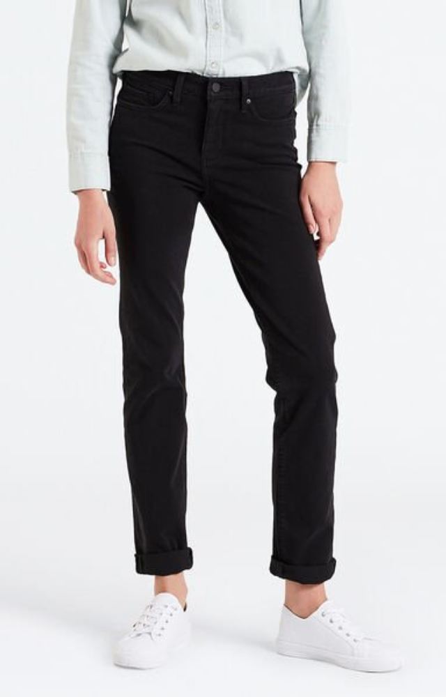 A person is standing against a plain white background wearing tummy-slimming black Levis 312 Shaping Slim Jeans, a light long-sleeve button-up shirt, and white sneakers. The jeans are rolled up slightly at the ankles. The person's hands are resting by their sides.