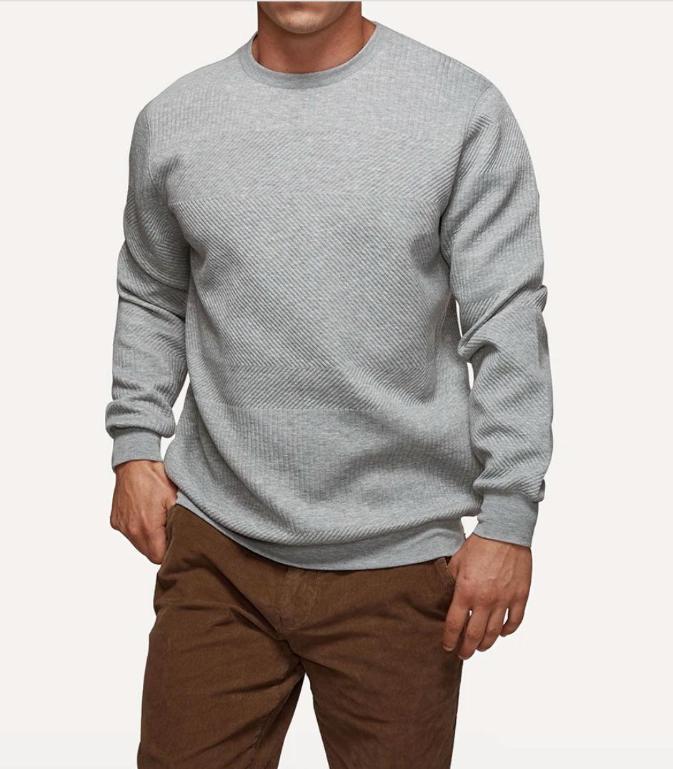 The individual is dressed in the Industrie The Robinson Crew, a light gray, long-sleeve casual jumper with a textured diagonal pattern, along with brown pants. They are situated against a plain, light background and their head is not visible in the image.
