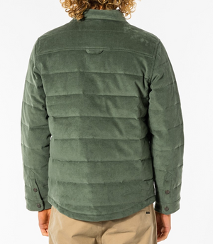 Rear view of a person wearing the forest green Rip Curl Convoy Anti Series Jacket with a stand-up collar. The water-resistant jacket features horizontal stitching and is paired with beige pants. The person's curly, shoulder-length hair stands out against the plain white background.