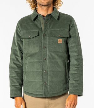 A person is wearing a Rip Curl Convoy Anti Series Jacket in forest green. The jacket, which is button-up and features a collar, has four front pockets, including two chest pockets with buttons. The individual with curly hair is paired with beige pants against a plain white background.