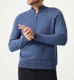 Wearing the Toorallie Grafton Half Zip Knit in blue, made from Australian Merino Wool, with long sleeves and black pants, a person stands against a plain light background with hands lightly clasped.