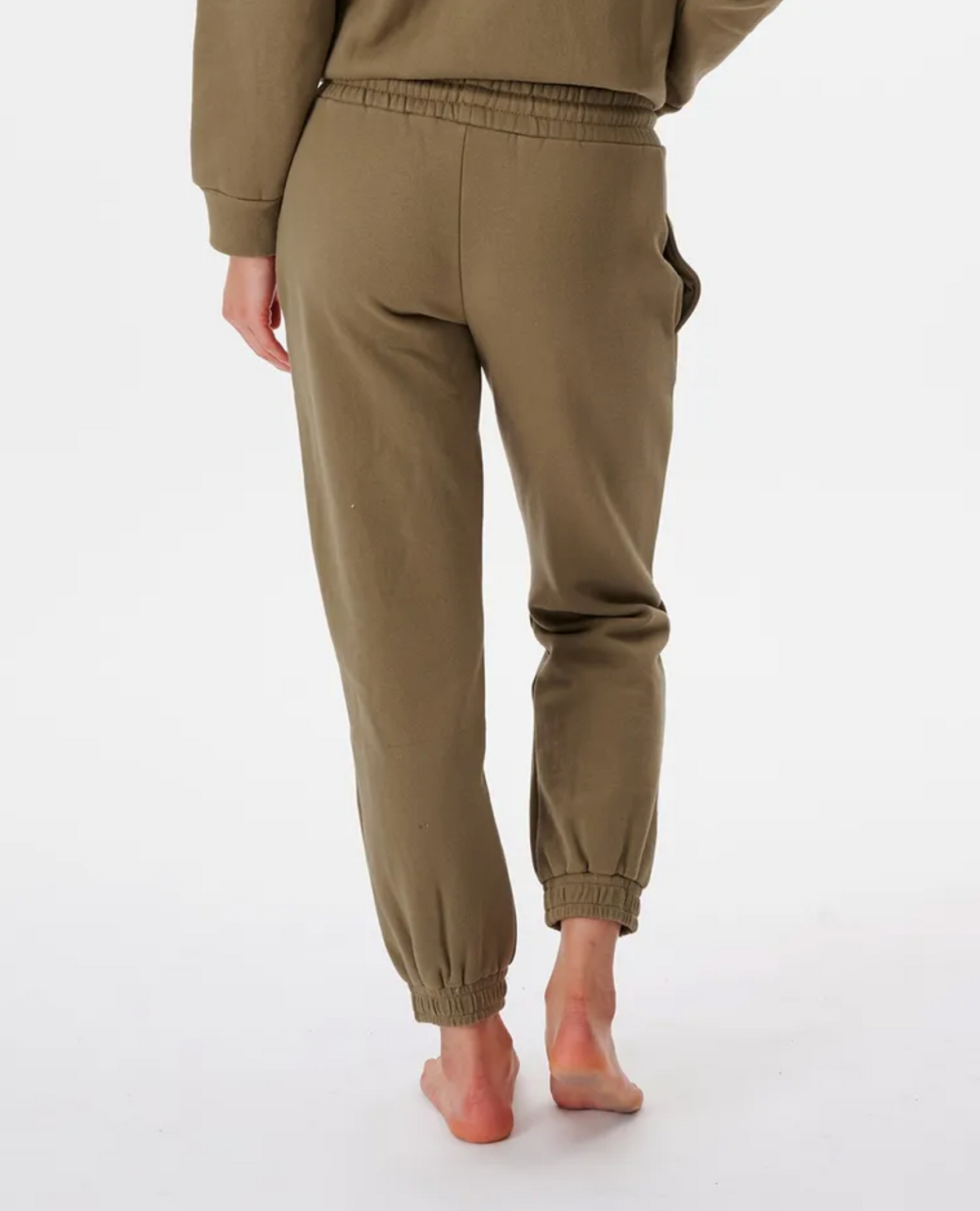 A person stands barefoot, facing away in olive green Rip Curl Premium Surf Trackpant with an elastic waist and cuffed style. The plain white background creates a studio-like setting, highlighting the casual elegance of the design.
