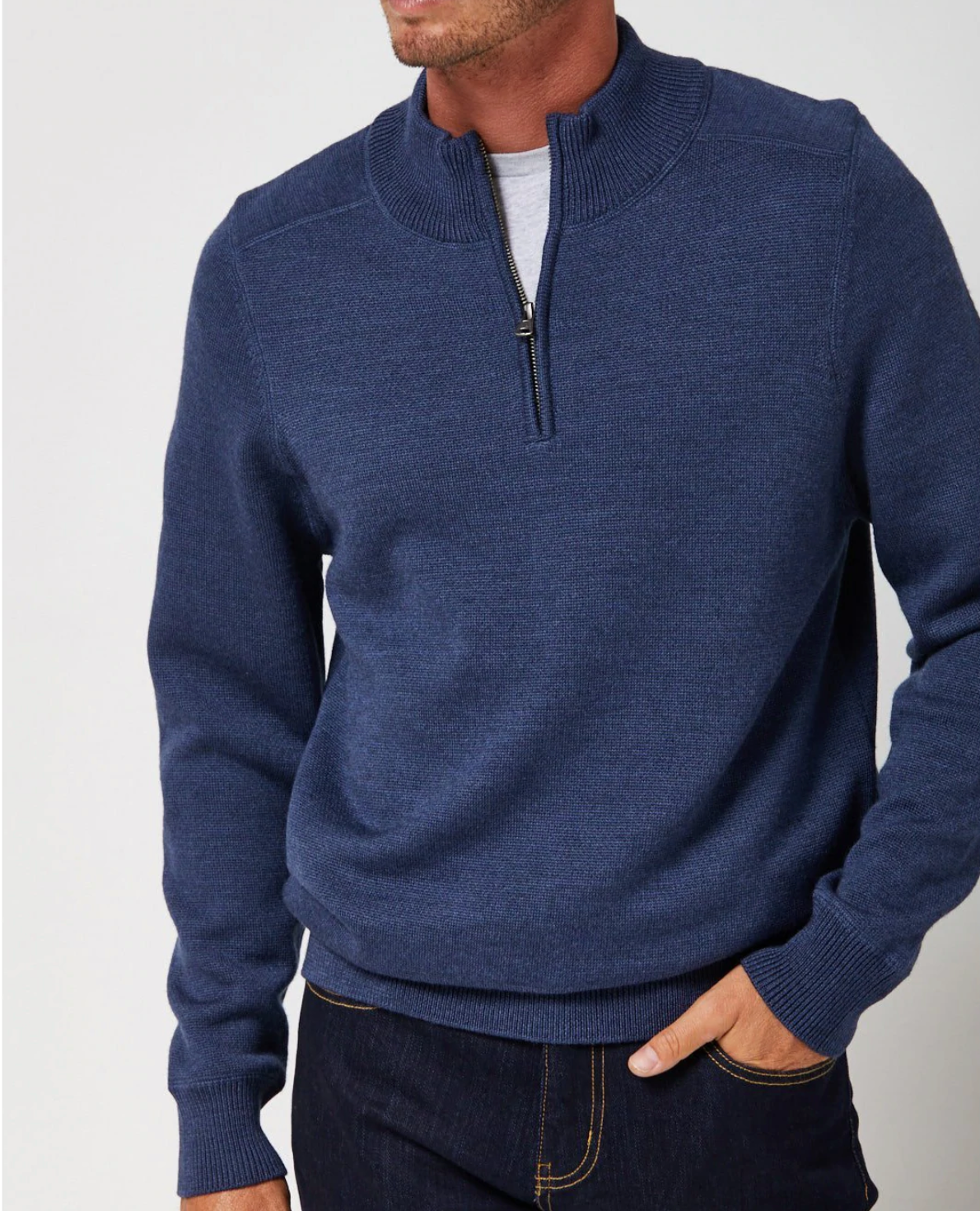 A person wearing a Toorallie Grafton Merino Polo in navy blue, paired with a light gray undershirt and dark blue jeans, stands with one hand in their pocket. The background is plain and light-colored, and the person's face is not visible.
