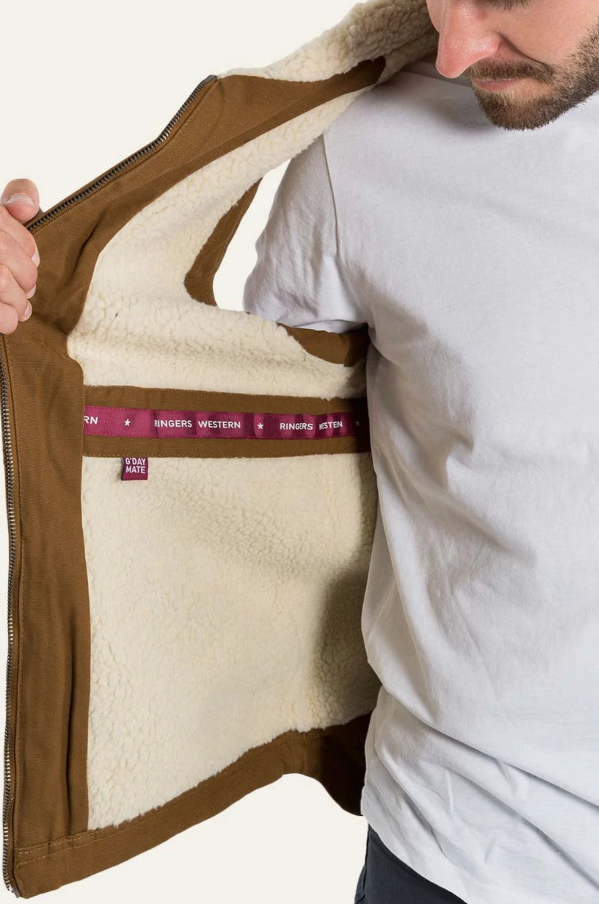 A person in a white shirt holds open the left side of a brown Ringers Western Macquarie Canvas Vest with sherpa-lined fleece, featuring maroon tape with white text inside. The person's face is partially visible, looking down at the vest.