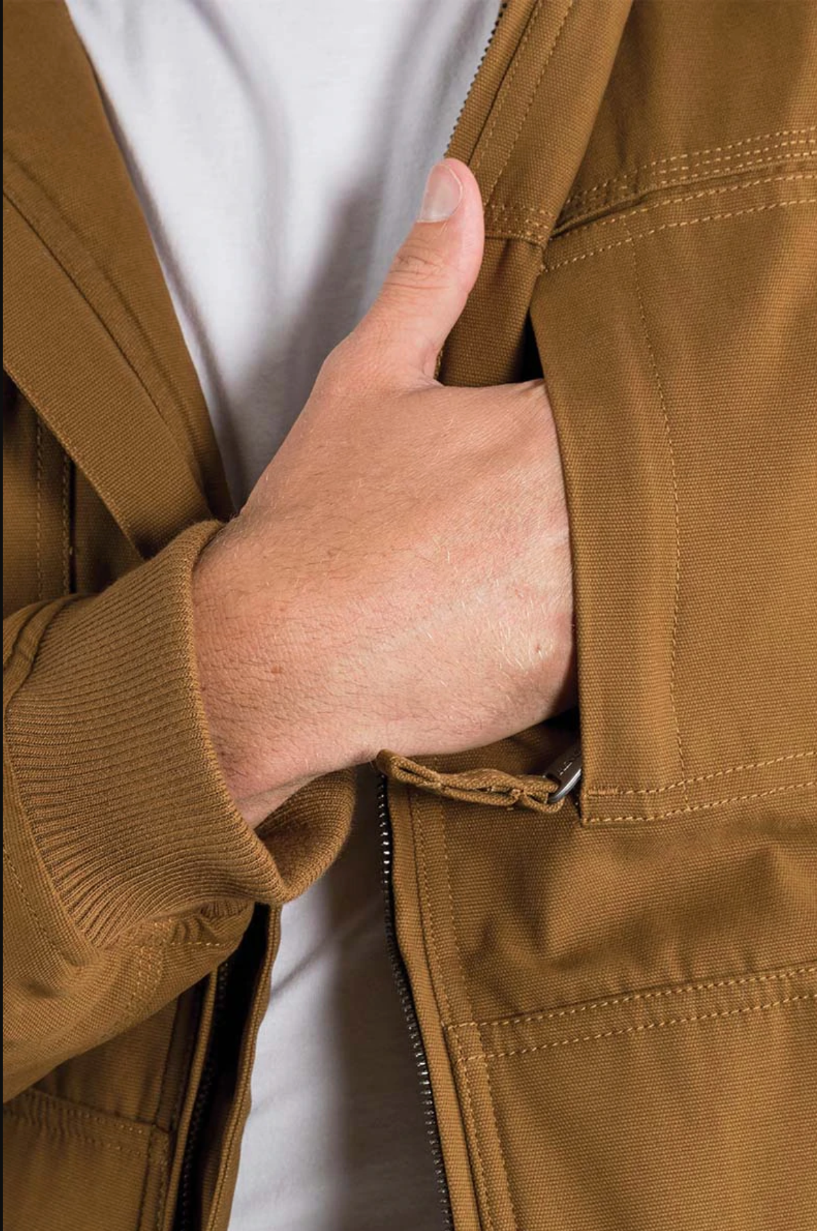 A person is using their left hand to pull down the zipper of a brown Ringers Western Kalgoorlie Long Canvas Jacket, partially revealing the white shirt underneath, with focus on the hand and jacket's zipper area.