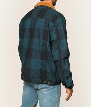 A person wearing a stylish Ringers Western Milawa Corduroy Jacket, featuring a blue and black checkered pattern and a brown collar, is standing with their back to the camera. The individual complements this comfortable cotton corduroy jacket with light blue jeans.