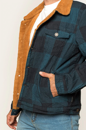 A person wearing the stylish Ringers Western Milawa Corduroy Jacket, characterized by its blue plaid design and brown sherpa lining, with a white T-shirt underneath, has one hand in their jean pocket while standing against a neutral background. This comfortable jacket offers both fashion and warmth, making it an ideal choice for any casual outing.