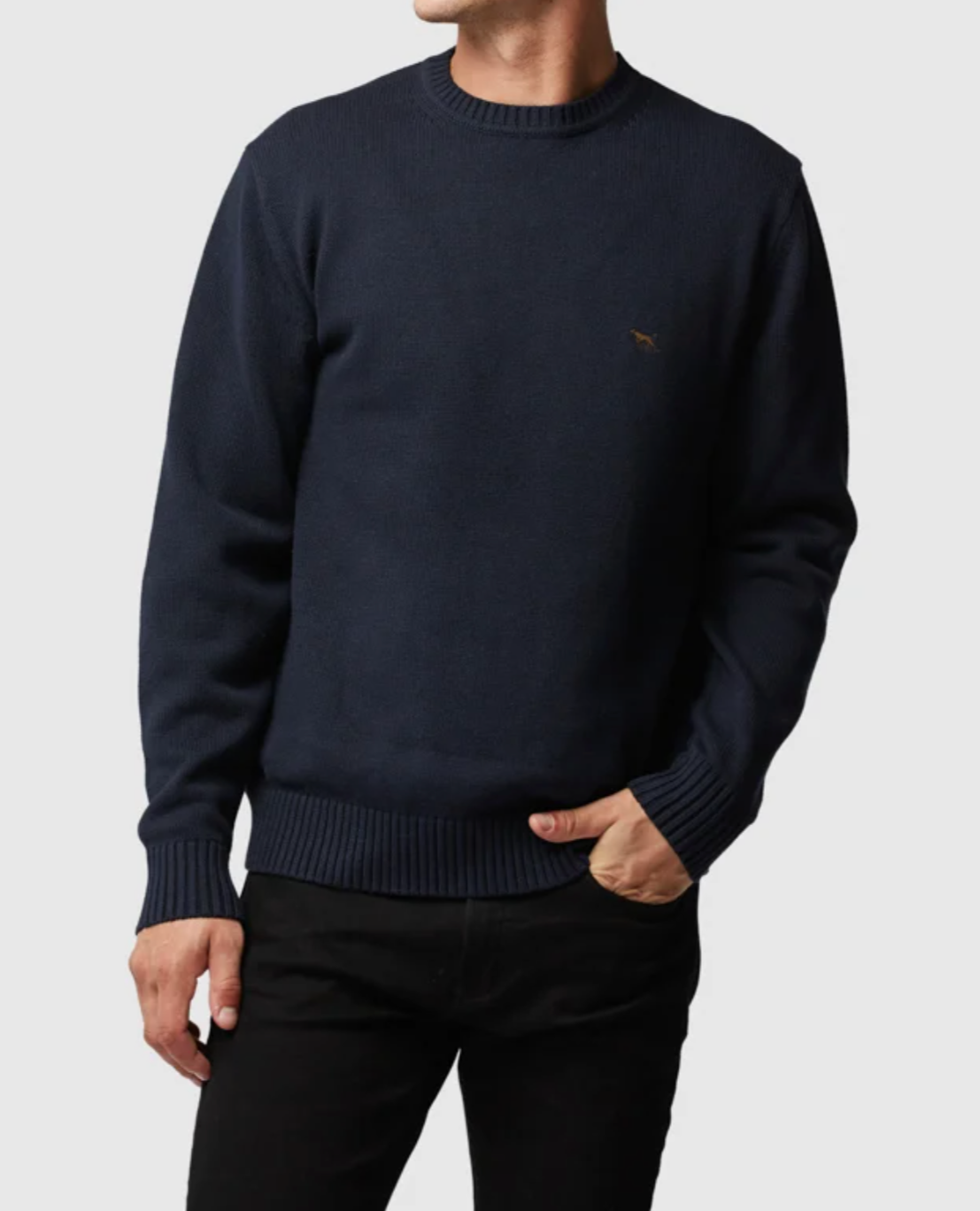 A person wearing the stylish Rodd & Gunn Gunn Knit sweater in dark navy blue, featuring a small embroidered logo on the left chest. The 100% cotton sweater boasts a ribbed crew neckline, as well as ribbed cuffs and hem. The person has one hand in their pocket and is dressed in dark-colored pants against a plain background.