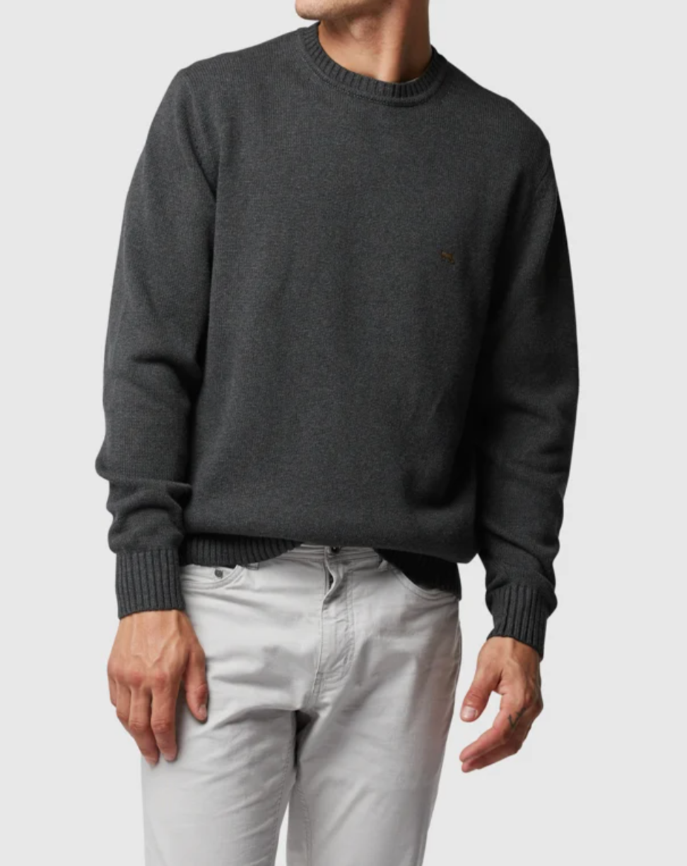 A person is wearing a dark gray, long-sleeve Rodd & Gunn Gunn Knit sweater and light gray pants. The comfortable sweater, made from 100% cotton, complements their relaxed pose with one hand slightly in their pocket and the other by their side against a plain gray background. Their head is partially out of the frame.