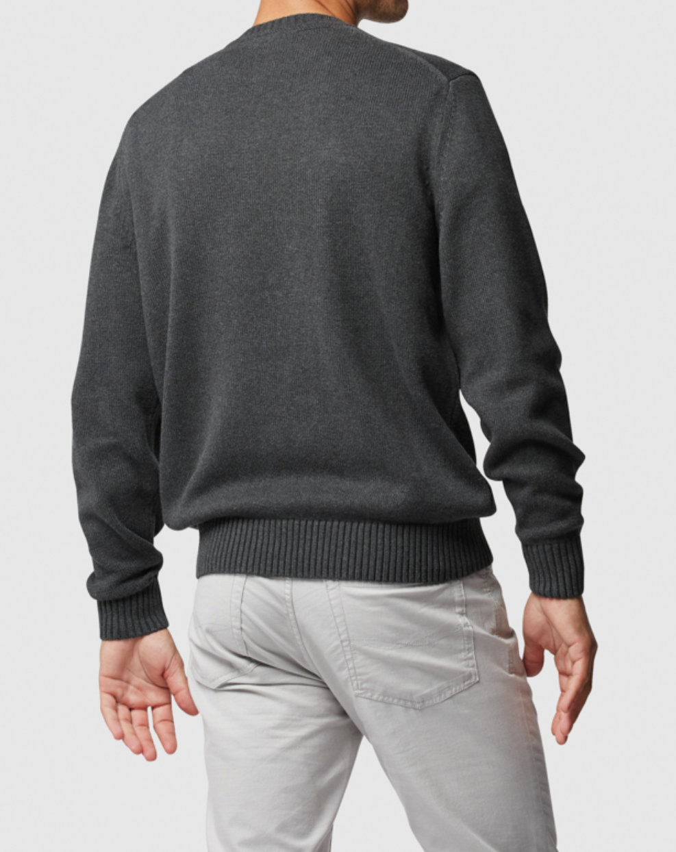 A man is seen from behind wearing a dark gray Rodd & Gunn Gunn Knit sweater, crafted from 100% cotton, paired with light gray pants against a simple backdrop.