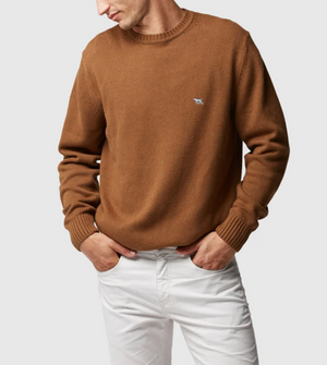 A man wearing a stylish Rodd & Gunn Gunn Knit brown sweater, made of 100% cotton, with a small embroidered logo on the chest and white pants stands against a plain background. His hands are in his pockets, and he's looking down to his right.