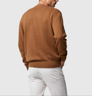 A person facing away from the camera, wearing a stylish Rodd & Gunn Gunn Knit brown sweater made of 100% cotton and white pants. The person has their left hand in their pocket, and the background is plain light grey.