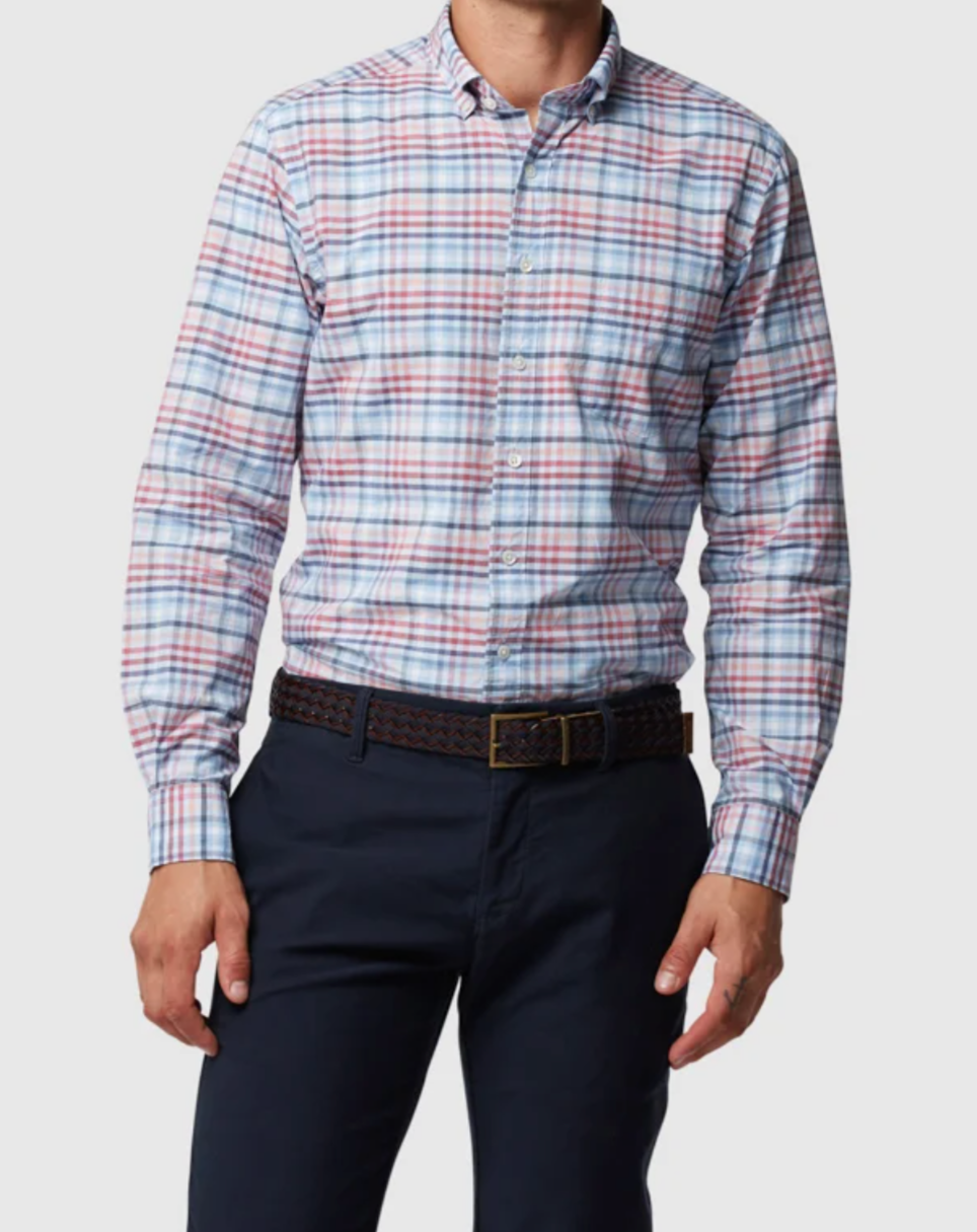 A person stands against a plain white background, wearing the stylish Rodd & Gunn Great Exhibition Bay Sports Fit Shirt, which is 100% cotton. The shirt features a white and red plaid design with intersecting blue and pink lines, paired with dark navy pants and a brown woven belt.