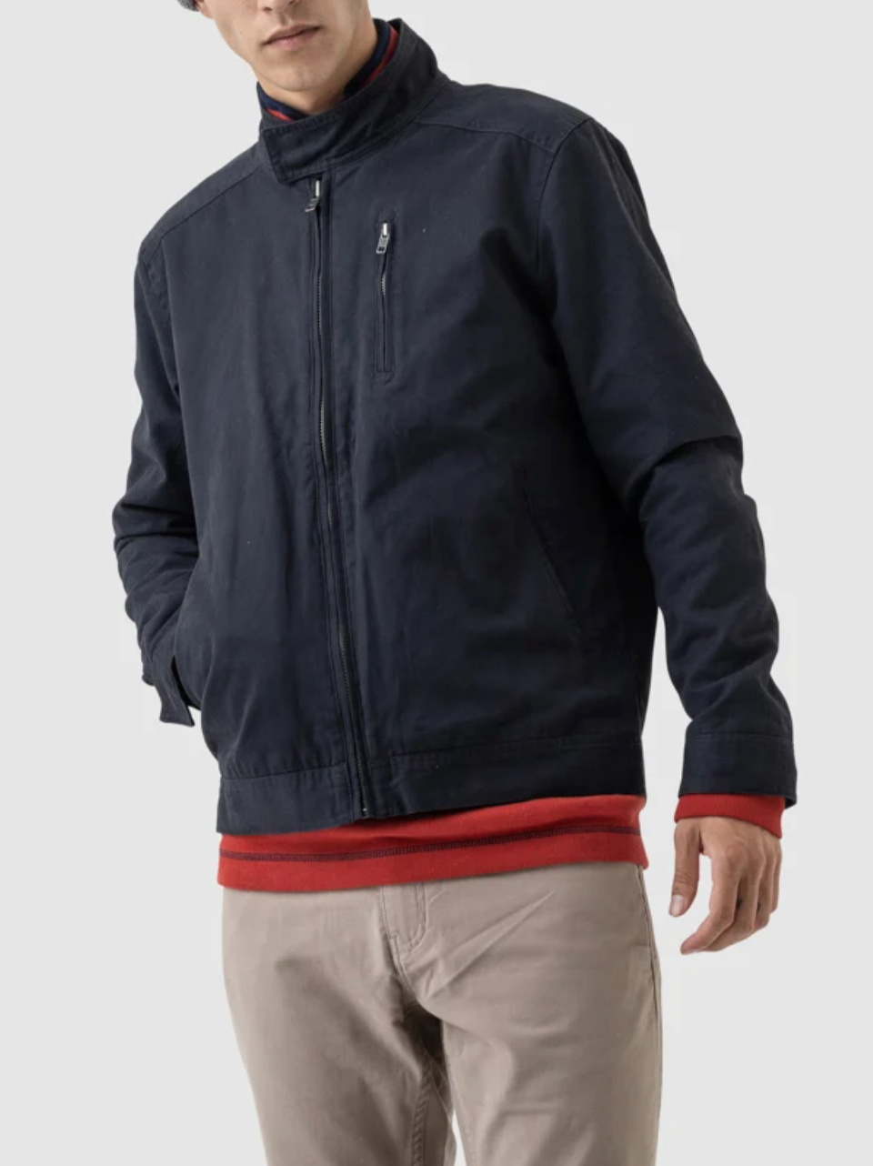 A person in a Rodd & Gunn Armitage Jacket, a stylish navy blue zip-up with adjustable cuffs made of Australian Cotton, over a red shirt and beige pants has their left hand in the pocket. The background is plain gray.