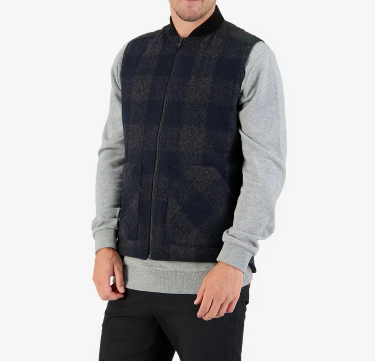 A person wears a Swanndri West Melton Wool Vest, featuring black and blue plaid with gray sleeves and a black collar, along with sleek black pants. The plain white background accentuates the ensemble's classic appeal as they stand with hands at their sides.