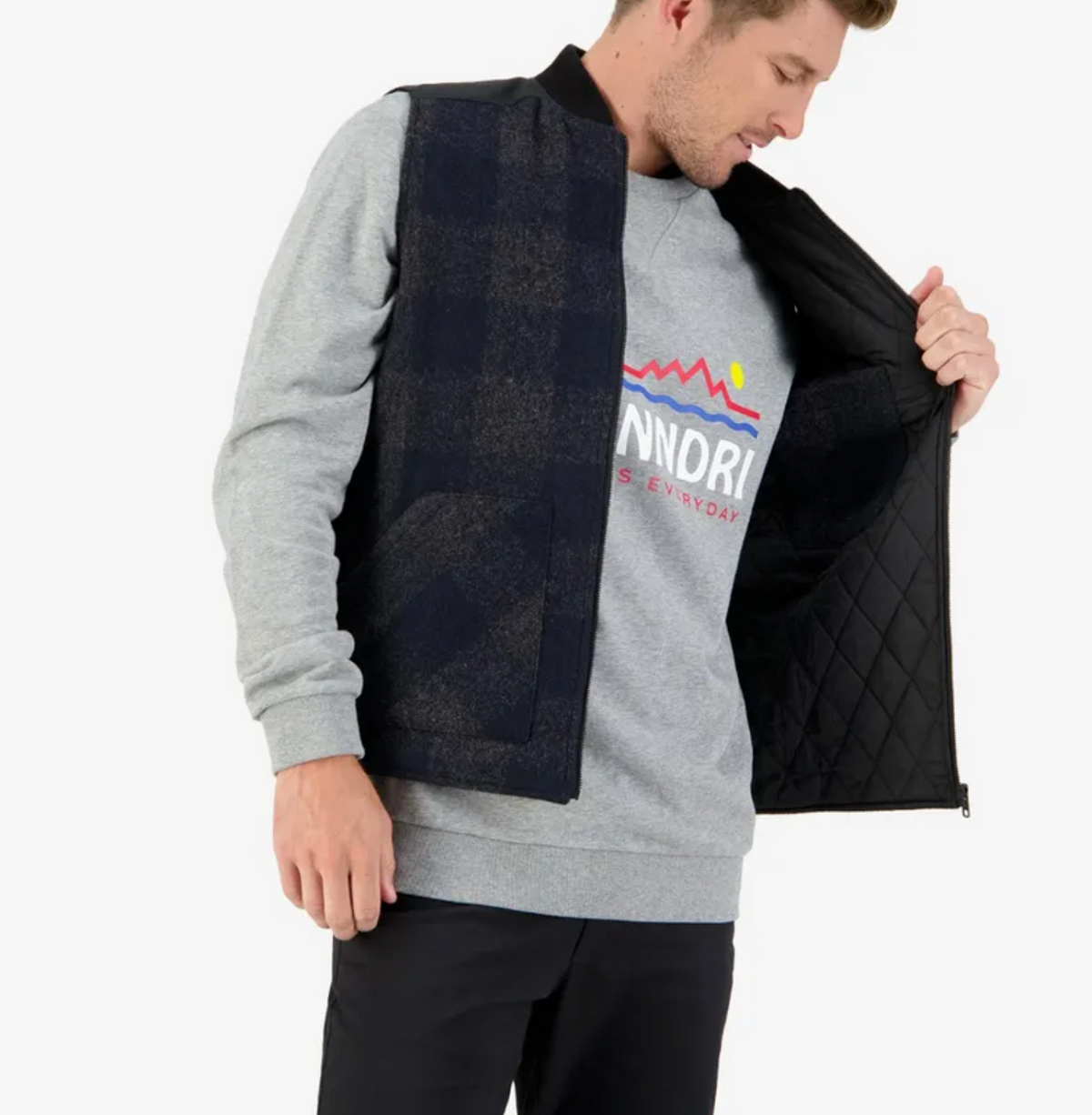 A man is wearing a gray sweatshirt with a colorful graphic, black pants, and is standing while unzipping an oilskin-panelled Swanndri West Melton Wool Vest with quilted lining. He looks down to the left, his right hand inside the open vest.