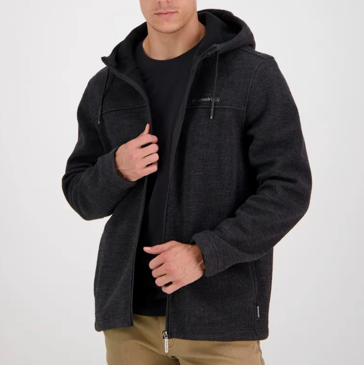 A man stands against a plain white background, partially unzipping his Swanndri Rifleman Wool Blend Hoody worn over a black shirt and khaki pants.