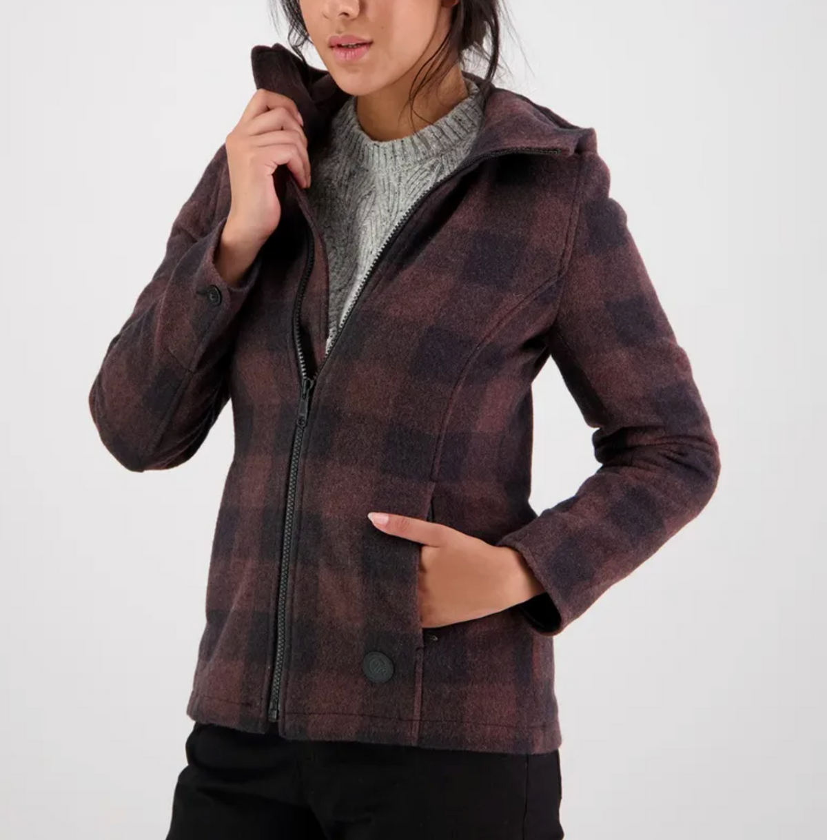 A woman wearing a checkered brown and black Swanndri Womens Seattle Hoody over a gray knit sweater stands with one hand in her pocket and the other adjusting her collar. She is posing against a plain white background, showcasing the contoured fit of her stylish outerwear.