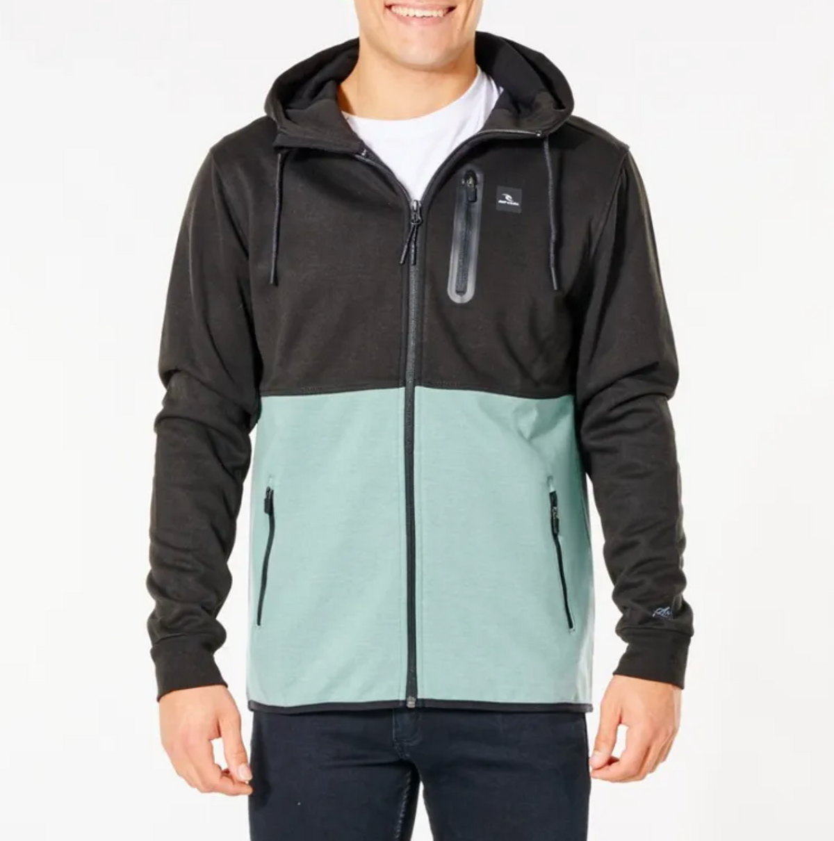 A person is wearing a Rip Curl Anti Series Departed Zip Thru hoodie, designed with a two-toned color scheme featuring a black upper section and a light green lower section. This Durable Water Resistant (DWR) hoodie includes a front zipper, two zippered pockets, and a small chest pocket with a zipper. The person is also dressed in dark pants and has a white shirt underneath.