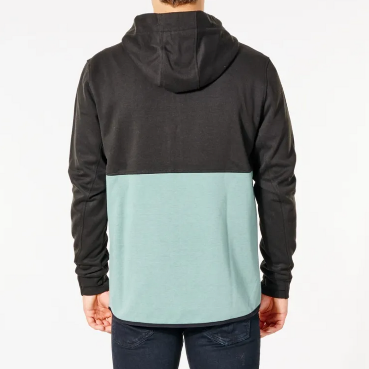 A person stands facing away from the camera wearing a Rip Curl Anti Series Departed Zip Thru two-tone hoodie. The top half of the hoodie is black while the bottom half is a light teal color. The person is also wearing dark blue jeans, completing their stylish winter ensemble with durable water resistance (DWR).