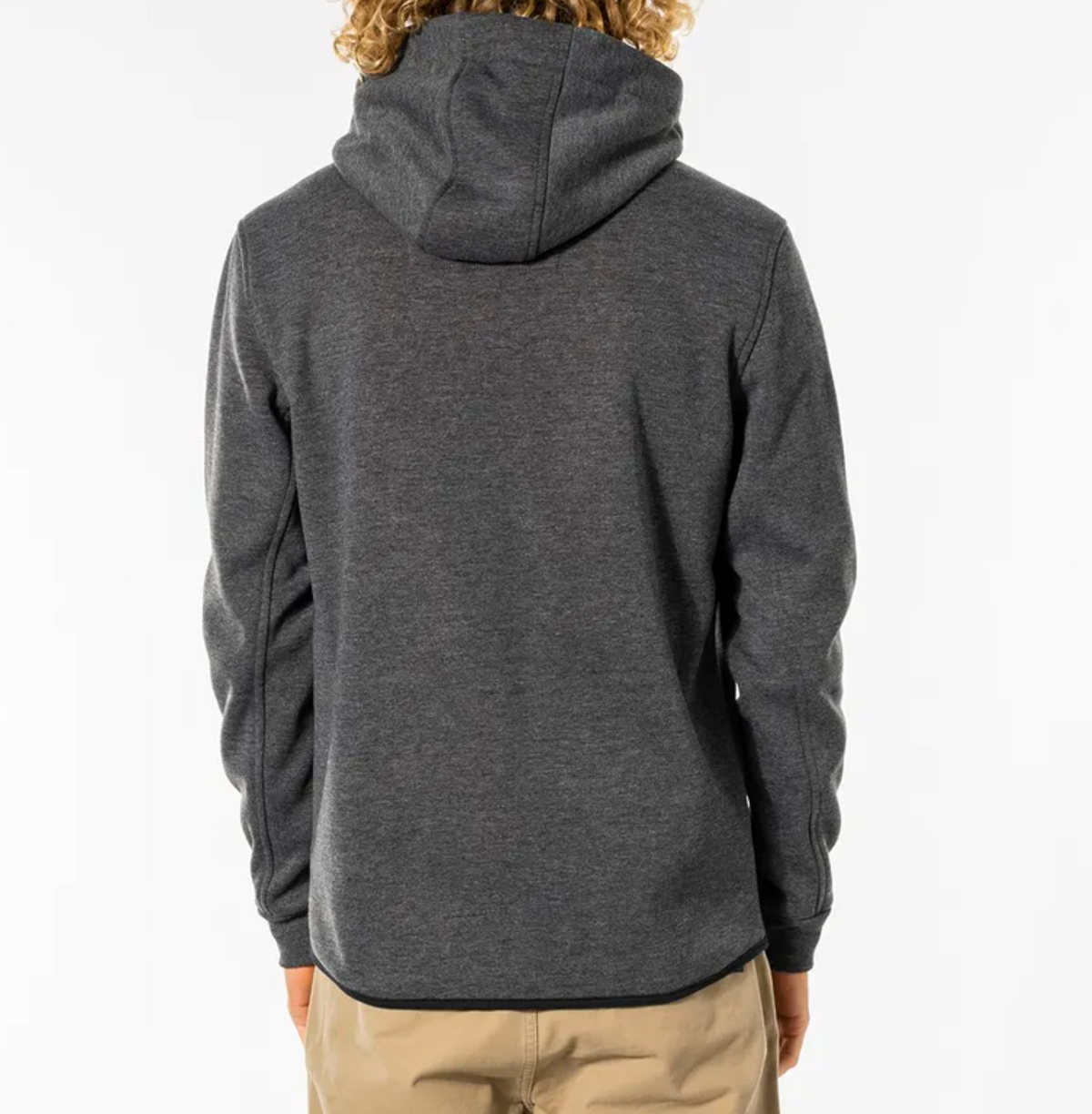 Back view of a person with long curly hair wearing a dark gray Rip Curl Anti Series Departed Zip Thru hooded sweatshirt and beige pants, standing against a plain white background.