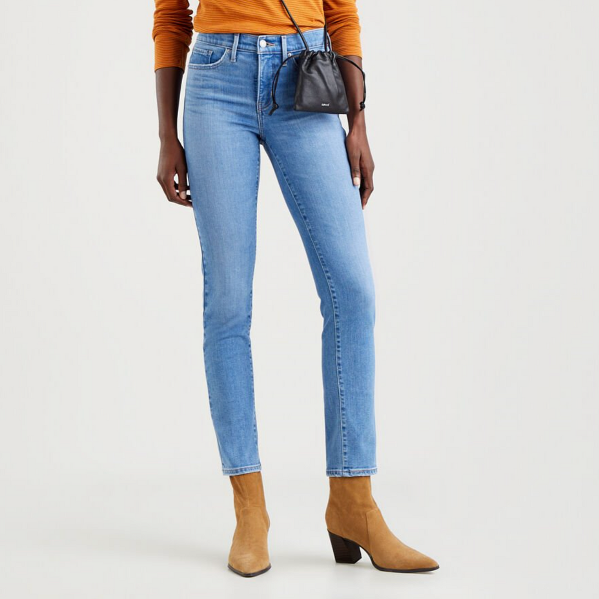 A person stands wearing light blue Levi's 312 Shaping Slim Jeans, featuring a tummy-slimming panel, paired with tan ankle boots with a low heel. They have a small black purse slung over their shoulder and are also wearing a burnt orange long-sleeve top. The background is plain white.